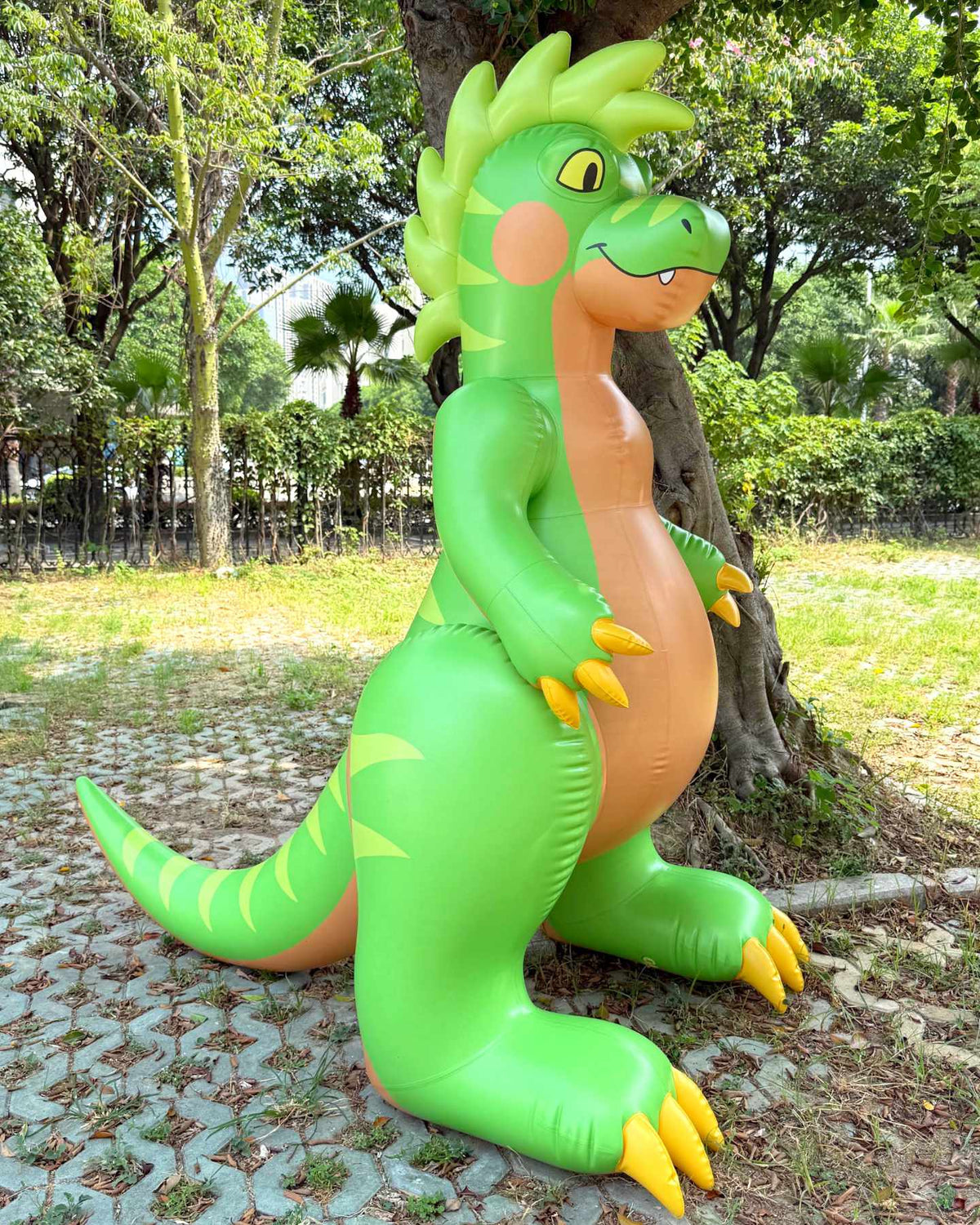 Raptary is a green inflatable dinosaur toy. Made by Jet Creations and Rapatary. Sold by SqueakClub. This photo shows Raptary outdoors next to a tree. Raptary is 6 feet tall. It is green with a tan belly, yellow claws, and wavy hair on its head. It stands on two feet with its tail on the ground.